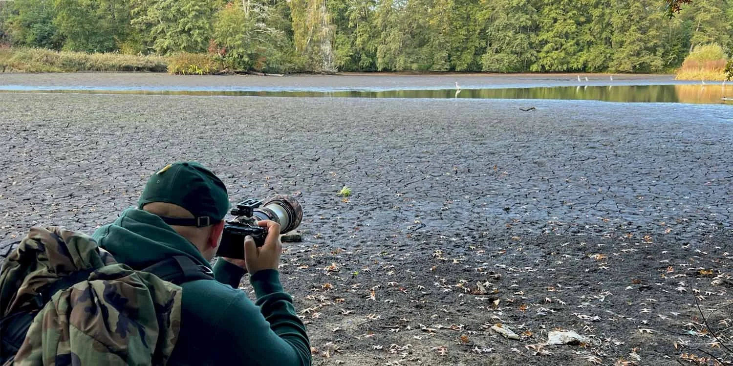 Fotograf mit Telekamera liegt auf rissigem, trockenem Seegrund und fotografiert Richtung Restwasser mit weißen Vögeln in der Ferne.
