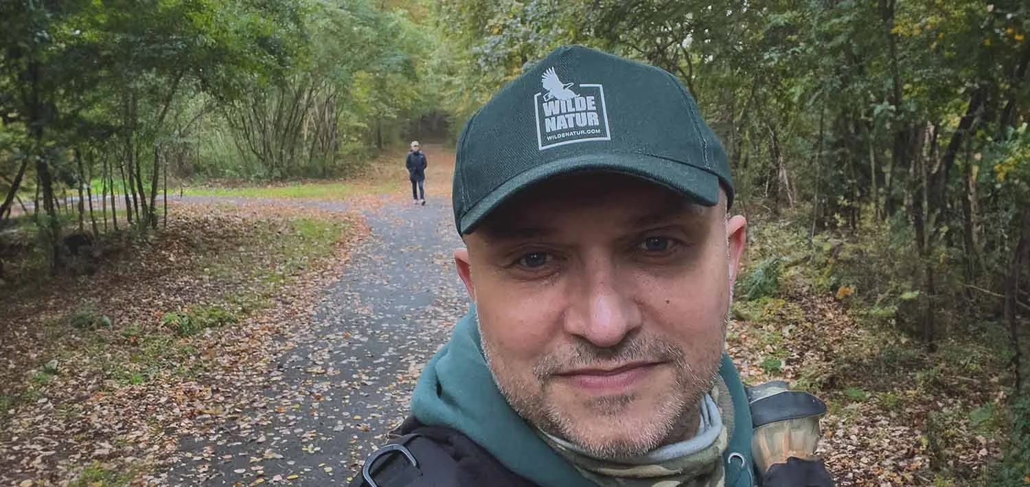 Selfie von Markus Nilles mit grüner WildeNatur-Cap auf einem herbstlichen, laubbedeckten Waldweg.