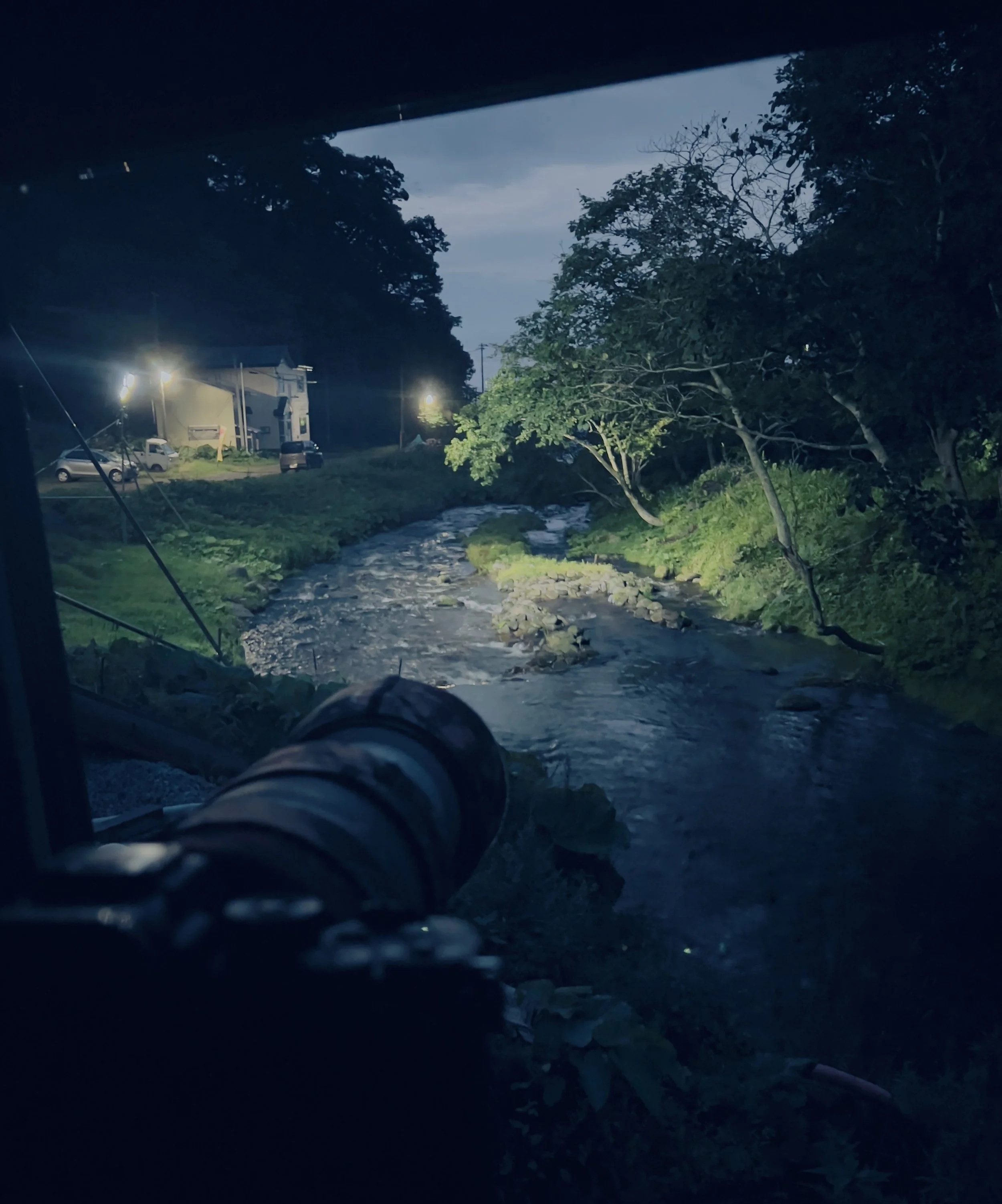 Telephoto lens aimed from vehicle window at a night-time stream, hide site for fish owl photography in Japan.