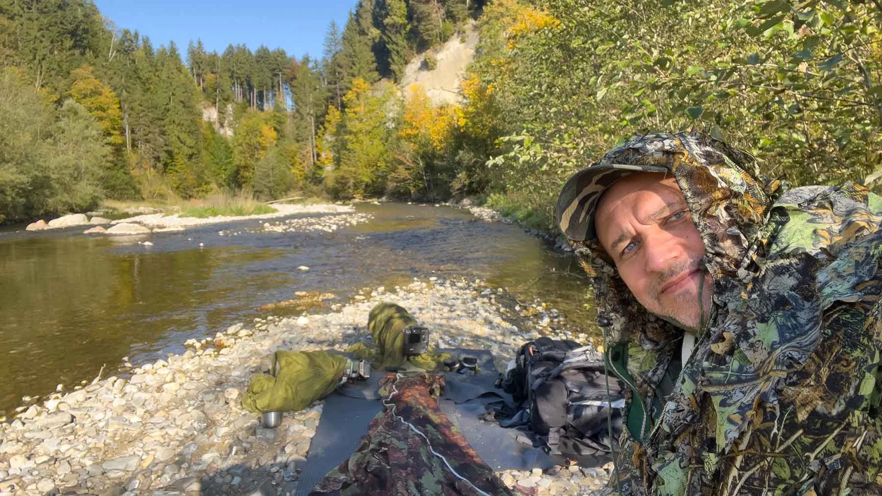 Fotograf Markus in Tarnkleidung am Kiesufer eines Flusses im Allgäu.